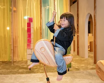 キッズスペースで、ブランコのような木製の遊具に乗って笑顔で遊ぶ女の子の画像です。活き活きとした表情から、遊びを心から楽しんでいる様子が伝わってきます。